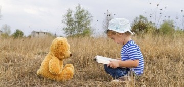 Ein Junge mit einem Buch und ein Teddybär sitzen sich gegenüber auf der Wiese